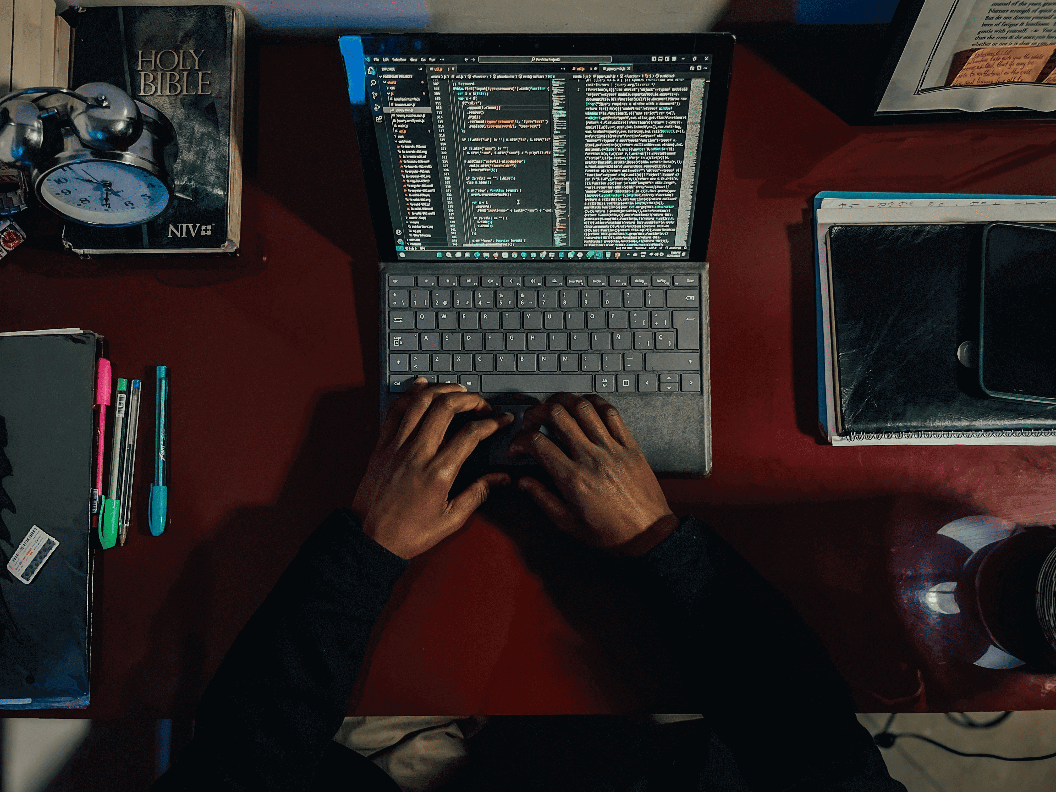 Рабочий стол крупным планом: на нем стоит ноутбук с кодом на экране, а руки человека печатают код на клавиатуре.