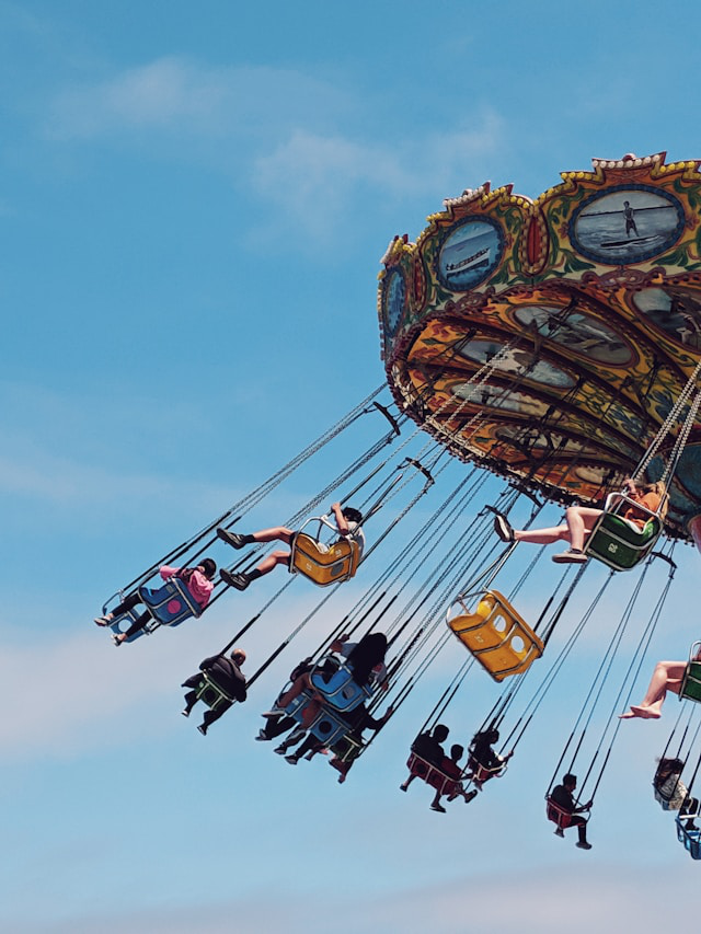 Люди катаются на аттракционе цепочная карусель под ясным голубым небом. Карусель наклонена, сиденья с пассажирами раскачиваются наружу от центробежной силы.
